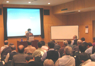 壇上の加部先生が大勢の受講者を前に話をしており、後方には大きなスクリーンとホワイトボードが設置されている、講座の様子の写真