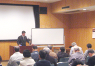 演台に立った土生田先生が受講者に話をしており、その横にはプレゼンテーション用のスクリーンとホワイトボードが設置されている、講座風景を撮影した写真