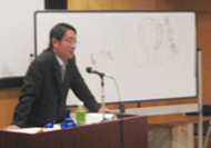 土生田先生が演台に立ってマイクに向かって話しており、後ろにあるホワイトボードには図や文字が書かれている様子を撮影した写真