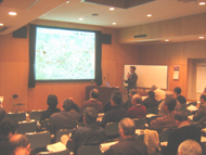 演壇に立つ柏木先生が大きなスクリーンに映し出された地図のような画像を指差して、着席した大勢の受講者に説明をしている、講座の様子の写真