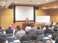 ホワイトボードの横に設置された大きなスクリーンを背に、演台に立った小川先生が、席に座った大勢の受講者に向かって話をしている、講座の様子の写真