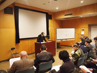 ホワイトボードの横に設置された大きなスクリーンを背にして、演台に立った久保田先生が、着席したたくさんの受講者に向かって話をしている、講義の様子を撮影した写真