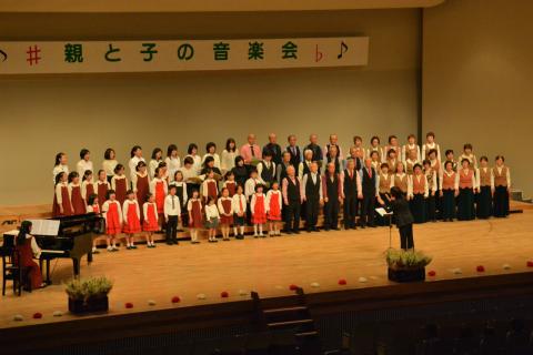 「親と子の音楽会」の横断幕が掲げられたステージ上に数十人の老若男女が正装した衣装で合唱しており、ピアノの伴奏者と舞台中央手前で指揮が指揮をしている様子を客席側から捉えた写真