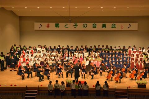 「親と子の音楽会」と書かれた横断幕の下、多数の児童や生徒が合唱隊として舞台上に並び、手前ではオーケストラの演奏者が指揮者の方を向き、客席からのアングルでコンサートホールのステージを捉えた写真