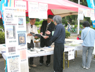 イベント会場の屋外ブースで、男性2人がテーブルに並んだ書籍を前に来場者と話しており、ブースの看板には書籍名と価格が書かれている販売の様子の写真
