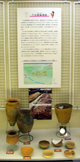 複数の土器が展示台に並べられ、背景の壁には下大槻峯遺跡に関する説明文と地図や航空写真が掲示されている写真