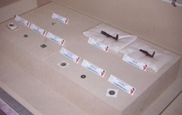 透明なケースの中で、白い布の上に置かれ、それぞれに説明書きの札が添えられている複数の古銭と、L字型の鉄片の展示物の写真