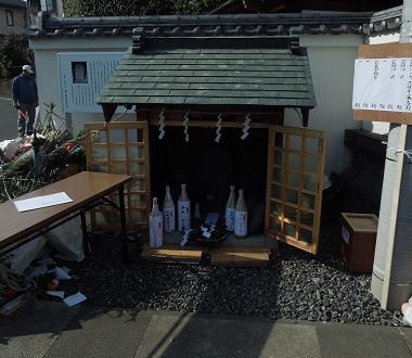 双体道祖神がまつられた小さな祠の格子状の扉が開き、中に複数の酒瓶などが供えらており、手前には神事用の長机が設置されている様子の写真