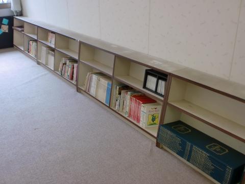 白い壁際に2段の書棚があり半分ほど本が入っていて、床はカーペット敷の児童室の写真