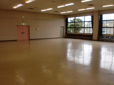 蛍光灯照明の天井、磨かれた茶色の床、左側に薄いピンクのドアと壁掛け時計、右側に黒い枠の大きな窓から自然光が差し込む広々とした多目的ホールの写真