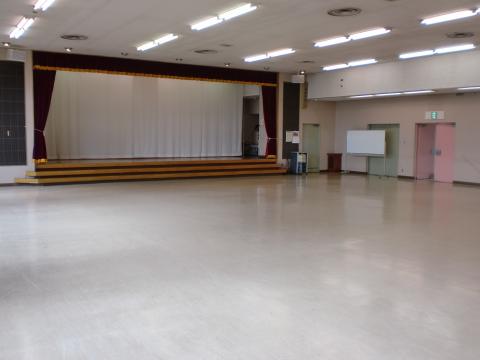 蛍光灯照明の白い天井、ベージュ色の磨かれた床、正面に木製階段があるステージと白い幕に赤いカーテン、右側に緑のドアとピンクのドアがある広々とした多目的ホールの写真