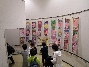 水彩画で顔を書いた画用紙二十数枚が壁面に展示されているのを大人数人が鑑賞している様子