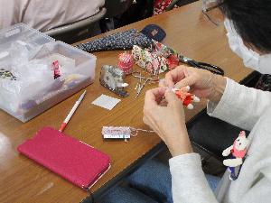 テーブル上に針仕事用の道具が置いてあり、大人が椅子に座って針仕事で小さな人形を作っている様子