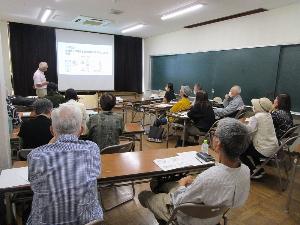 講師が資料をプロジェクターに映し説明しているのを10数名の受講者が聴いている。