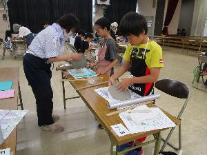 複数のテーブルそれぞれで、数人の小学生が粘土で工作をしているのを数人の大人が指導している様子