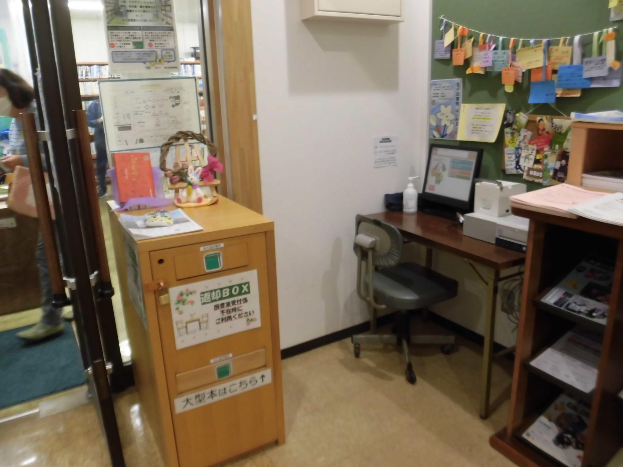 カウンターとパソコンが置かれた事務スペースに、返却ボックスや様々な掲示物が壁に貼られている図書室の入り口付近の写真