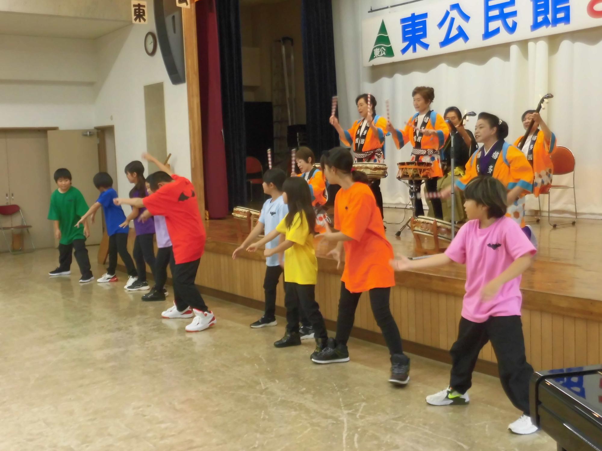 白い壁に「東公民館」と書かれた横断幕が掲げられたステージで、オレンジ色の衣装を着た複数の太鼓演奏者が後方に並び、前方では赤、緑、黄色、ピンクなどカラフルなTシャツを着た子どもたちが円を描いて踊っている写真