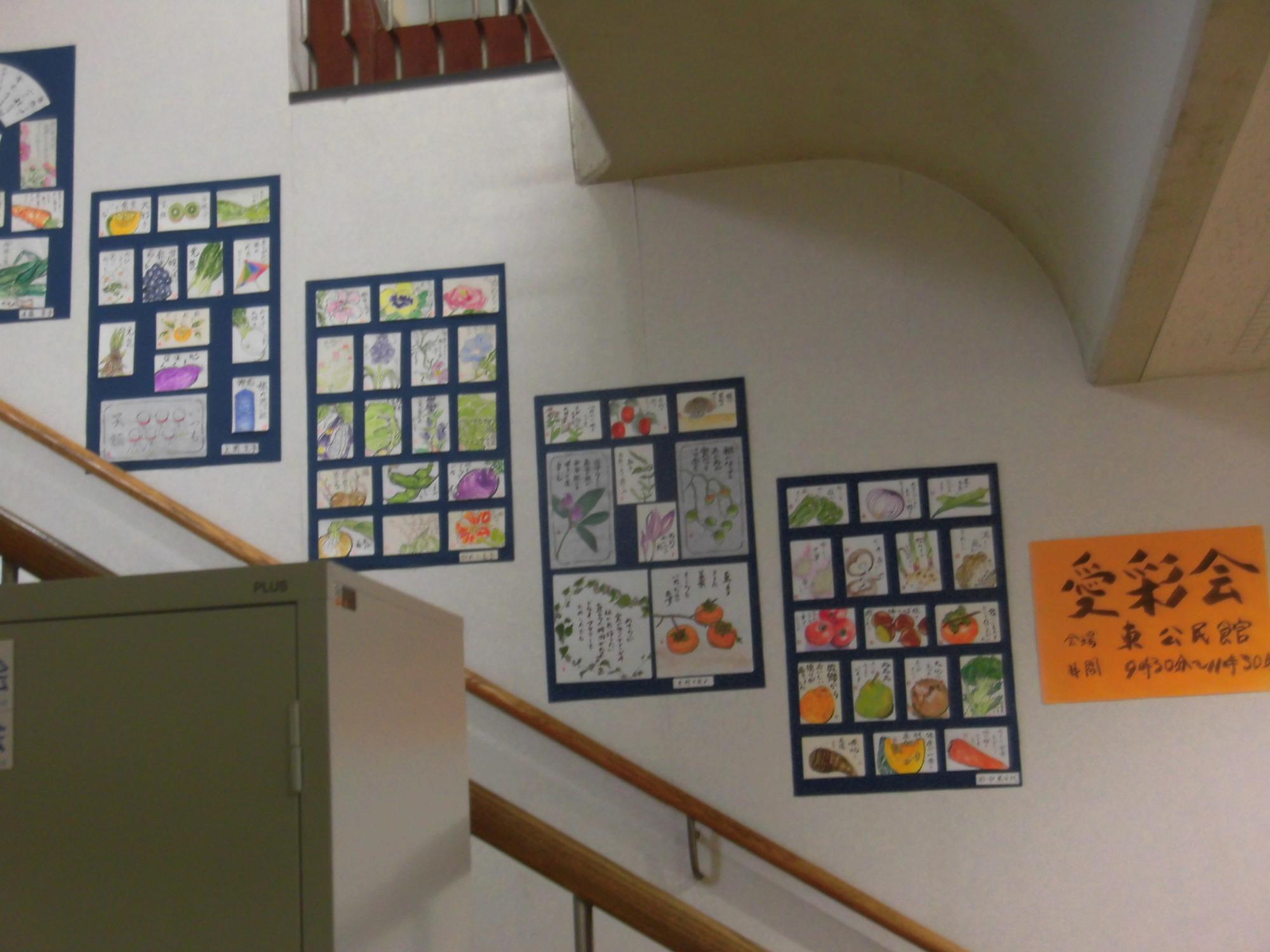白い壁に沿った階段の壁面に、野菜や果物を描いた複数のカラフルな絵手紙が紺色の台紙にまとめられて展示され、右端にオレンジ色の「絵彩会」と書かれたポスターが掲示されている写真
