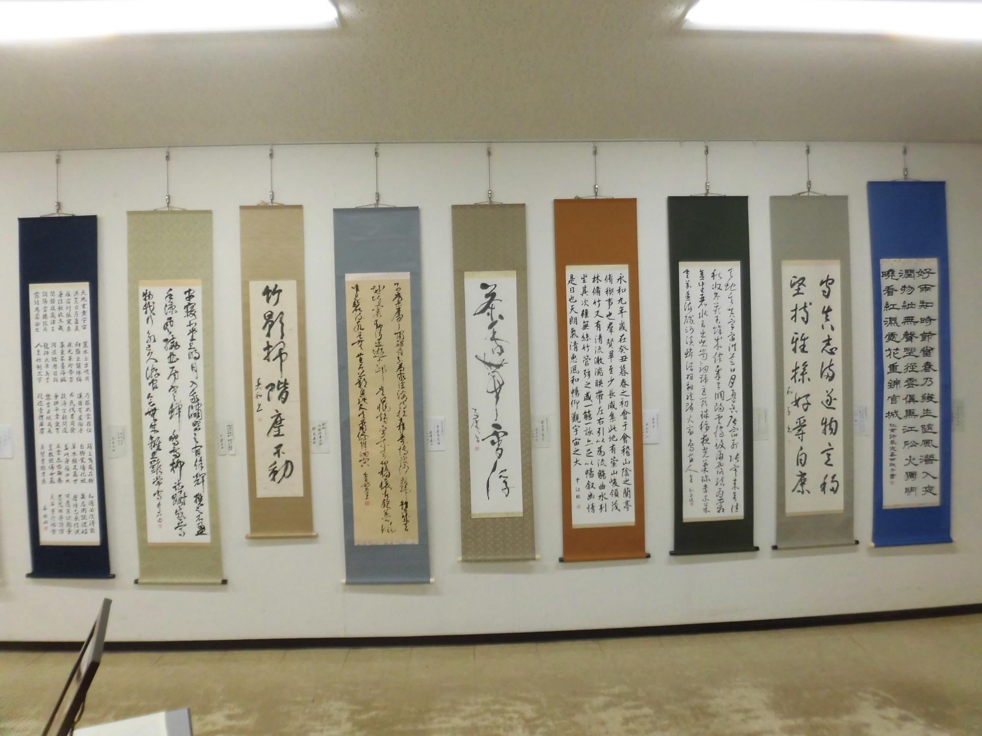 白い壁に紺、ベージュ、グレー、茶色、黒、青など様々な色の表装が施された9本の書道掛軸が横一列に吊るされて展示されている写真