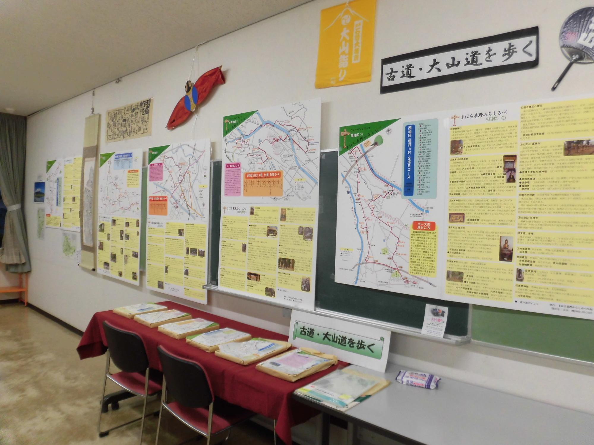 白い壁に「古道・大山道を歩く」と書かれた看板の下に、複数の地図やポスター、黄色い案内資料が掲示され、手前の赤い布を敷いたテーブルに複数の冊子や資料が並べられいる写真