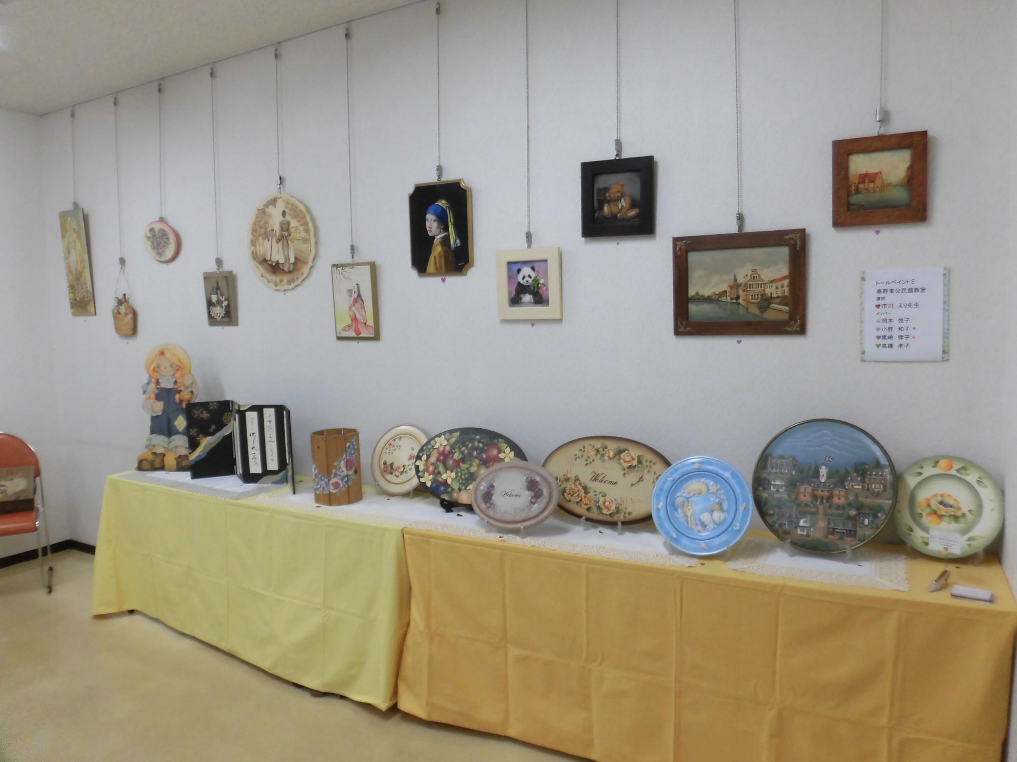 白い壁に複数の絵画や工芸作品が吊るされ、手前の黄色い布がかけられた長机の上に花や風景が描かれた円形や楕円形の装飾皿が並べられている写真