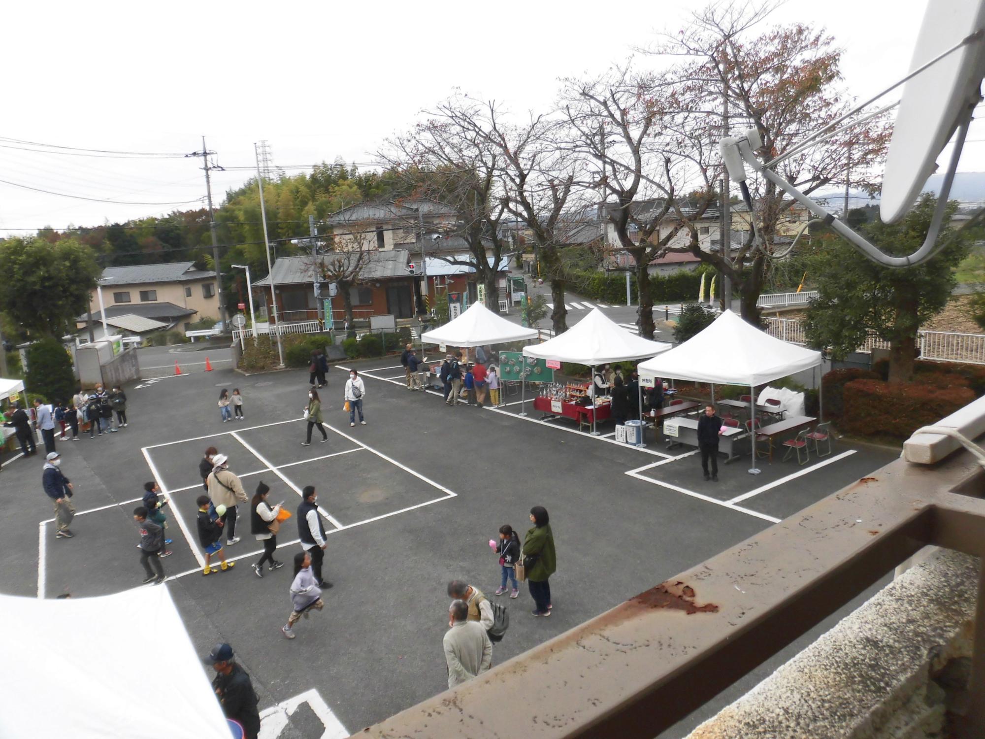 黒いアスファルトの駐車場に3つの白いテントが設置され、多数の参加者が歩き回ったりテントの周りに集まったりしている屋外イベントを高い位置から写した写真