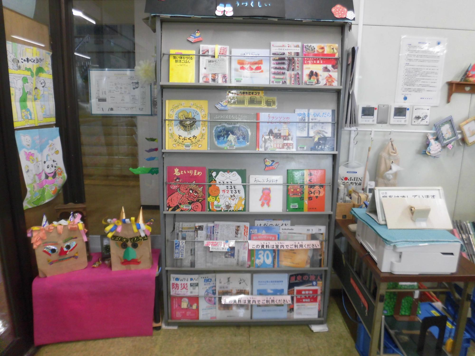 マガジンラックに雑誌や絵本が陳列され、その横の棚にピンクの布が敷かれ鬼のお面のような装飾が置かれている写真