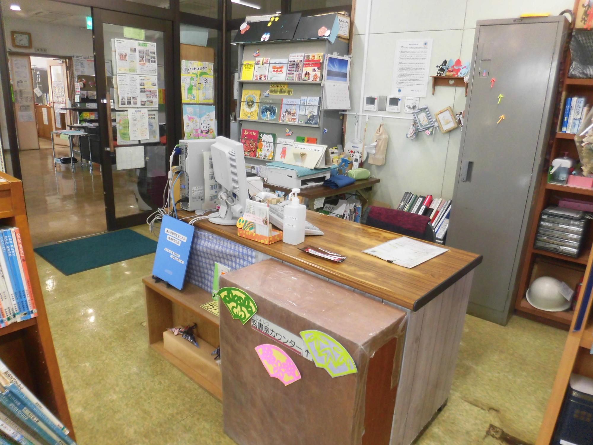 本の貸し出しや返却を行うカウンターが設置され、その上にはパソコンやプリンター、消毒液、掲示物があり、正面には「図書室カウンター」と書かれた棚が置かれている写真