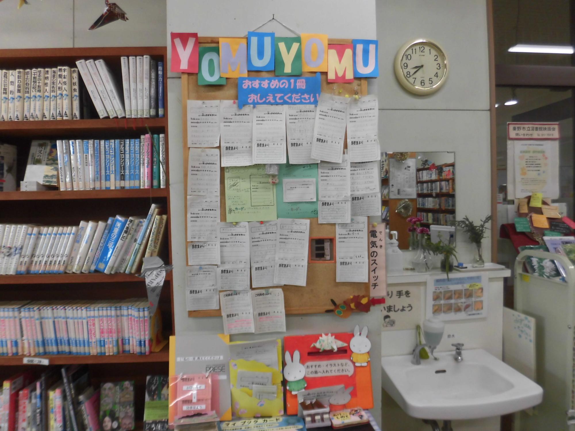 本棚の隣にある柱のコルクボードに「YOMUYOMU」と書かれたカラフルな文字や、おすすめの本を紹介する紙が多数貼り付けられている、図書室のコーナーの写真