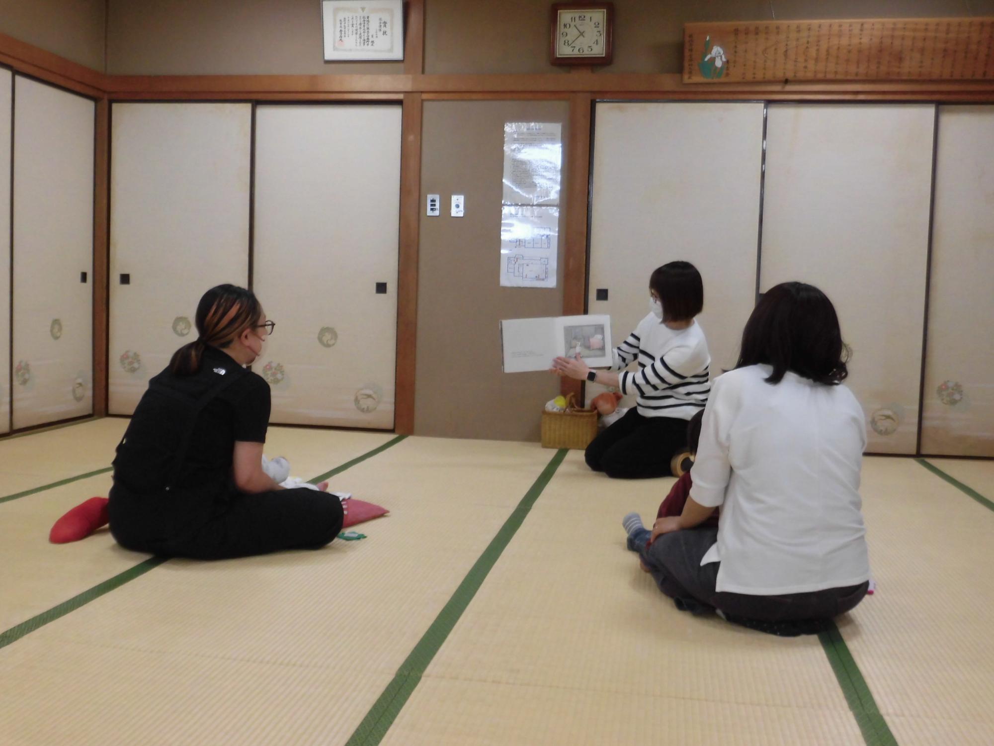和室で数人の女性が床に座り、そのうちの一人が他の人たちに絵本を読み聞かせている様子の写真