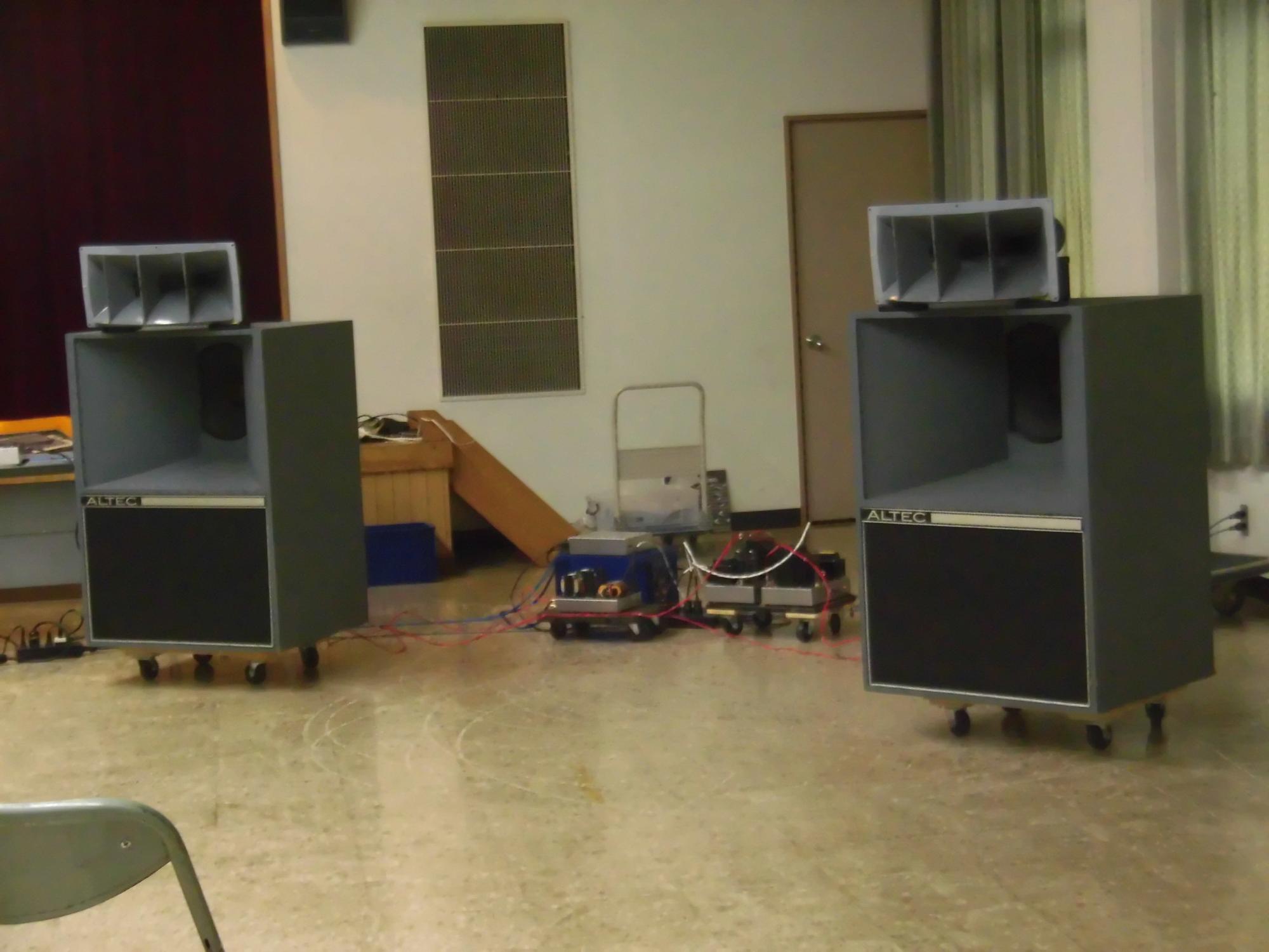 広い室内の中央に車輪付きの大型スピーカーが2台設置されており、それらがケーブルで接続されている音響設備の写真