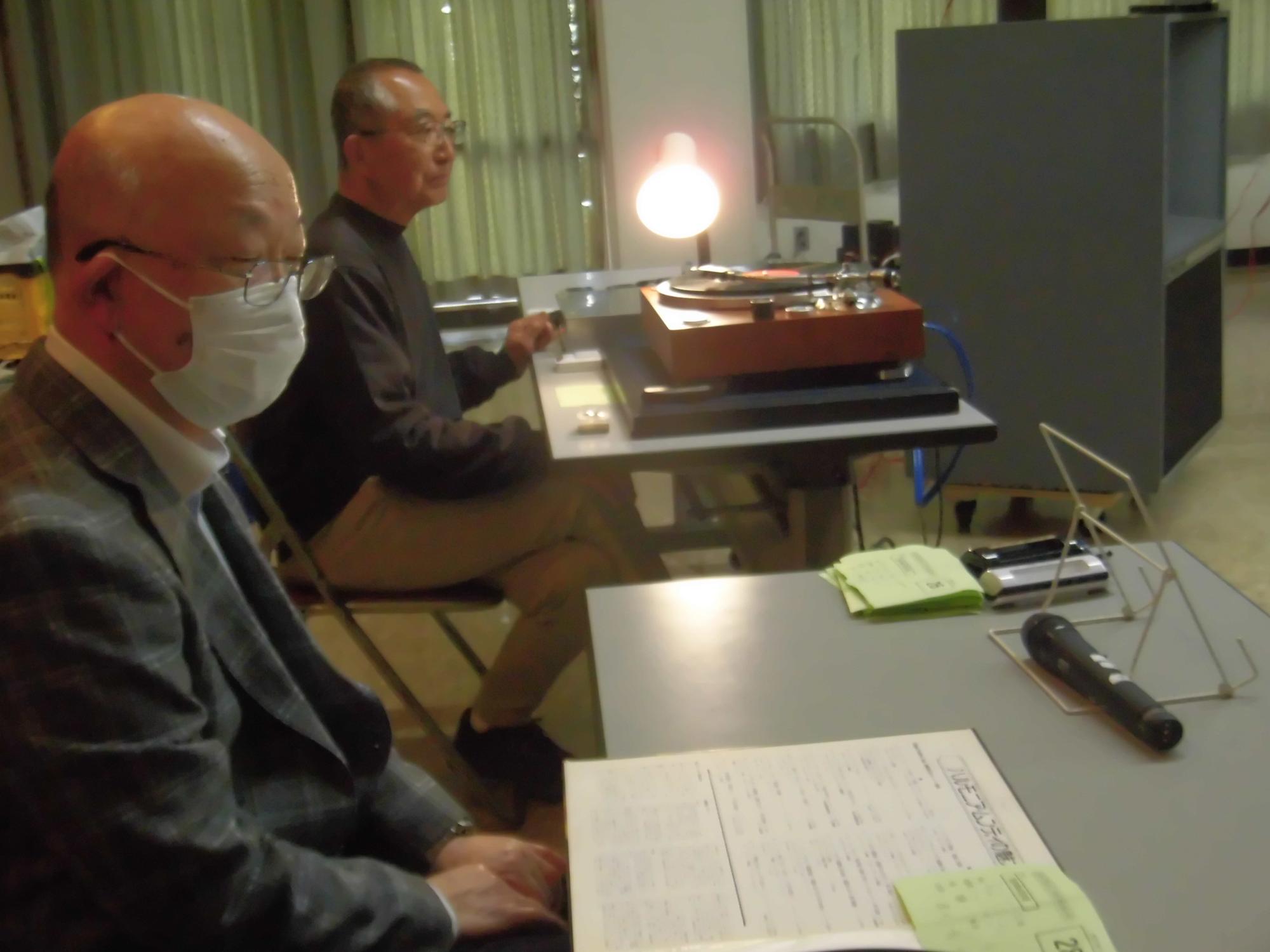 二人の男性がテーブルに向かい合い、一人がターンテーブルを操作し、もう一人がマイクと譜面が置かれたテーブルに座っている、レコード鑑賞会の様子の写真