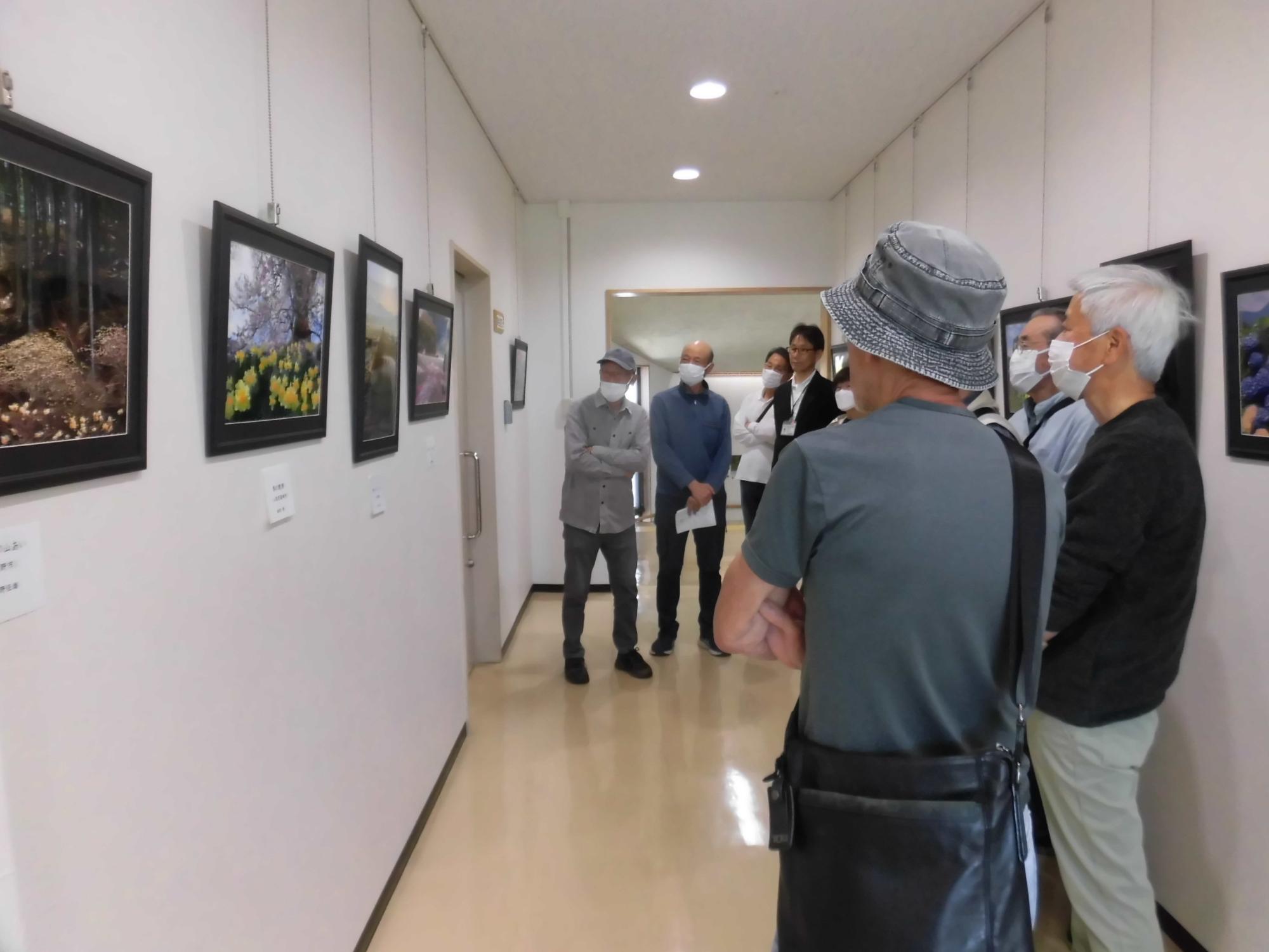 白壁の通路に写真が飾られており、マスクを着用した複数人の来場者がそれぞれの作品を鑑賞している、屋内で行われた写真展の様子を捉えた写真