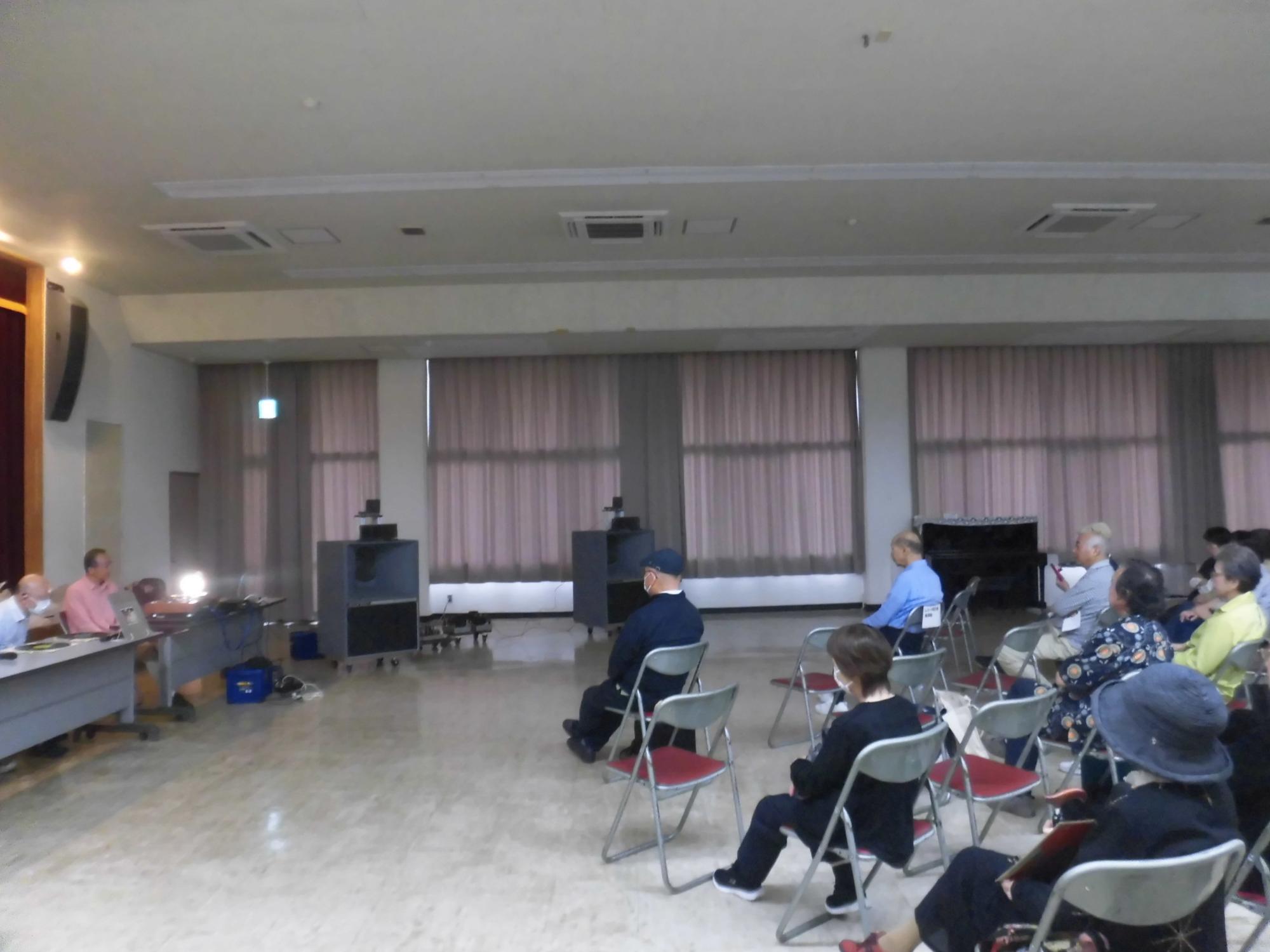 大きなホールで多数の参加者がパイプ椅子に座って、部屋の端で話す男性に注目している様子を写した写真