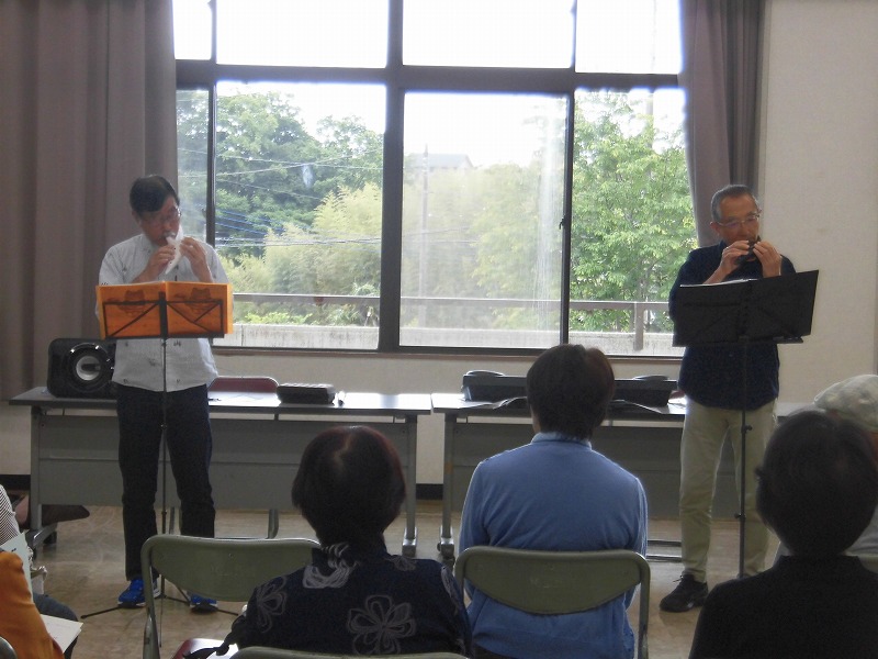 広い部屋の窓を背景に、2人の男性がそれぞれ譜面台の前に立ち、オカリナを演奏している様子を捉えた、観客を前にしたコンサートの写真