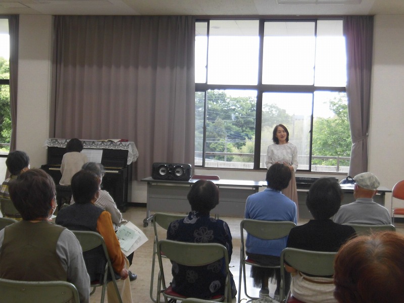 広々とした部屋で複数の参加者が椅子に座り、壇上の女性講師を見上げており、部屋の隅にはピアノが置かれている、音楽教室のような集まりの写真