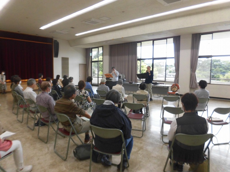 明るい光が差し込む広い部屋で、2人の男性が楽器を演奏し、パイプ椅子に座った多数の聴衆が鑑賞している、屋内で行われた音楽会の写真