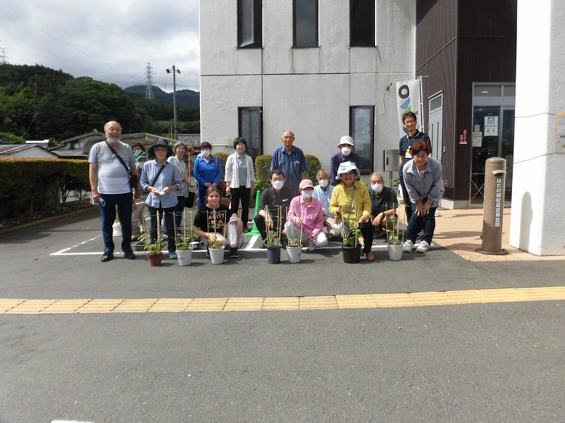 複数の高齢者が植木鉢に入った植物を囲んで並び、カメラに向かって笑顔で集合している写真