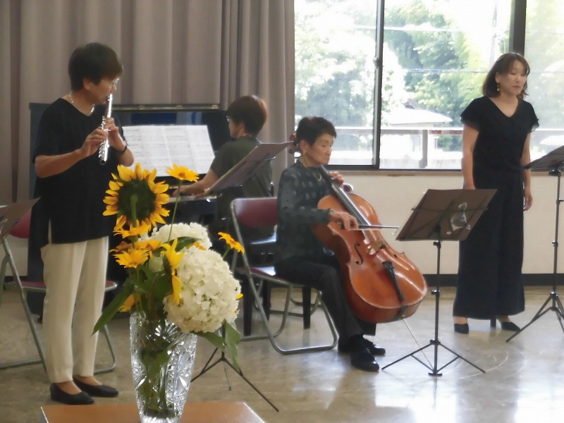 室内でフルート、チェロ、ピアノを演奏している三人の女性と、その隣に立って指揮をしている一人の女性を写した、東のお山のコンサートの写真