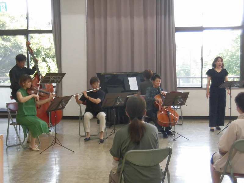 複数の弦楽器奏者がピアノとフルートの奏者と共に演奏しており、右端に立つ女性が口を大きく開けて歌っている、室内での小規模なクラシックコンサートの様子の写真