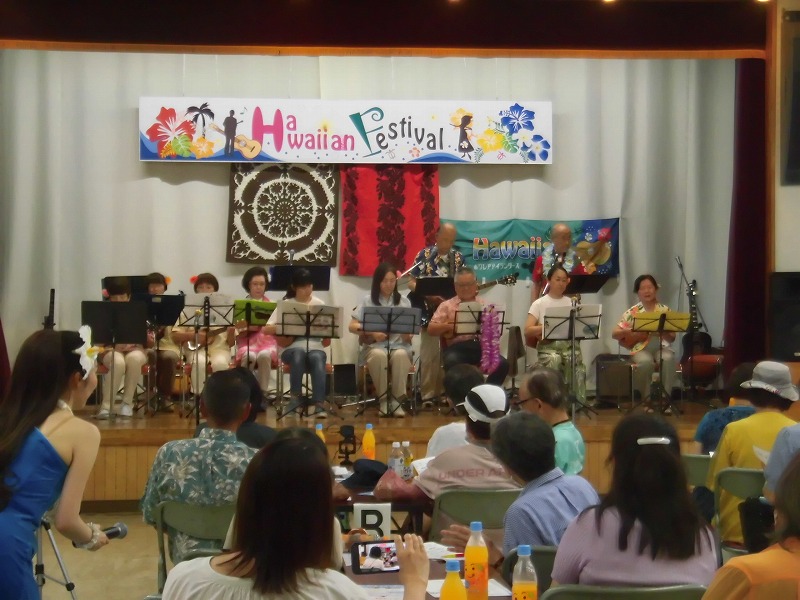 ステージの上で、楽器を持った複数の人々がハワイアン音楽を演奏しており、ステージ前では観客がそれを見ている、ハワイアンフェスティバルのライブ演奏風景を捉えた写真