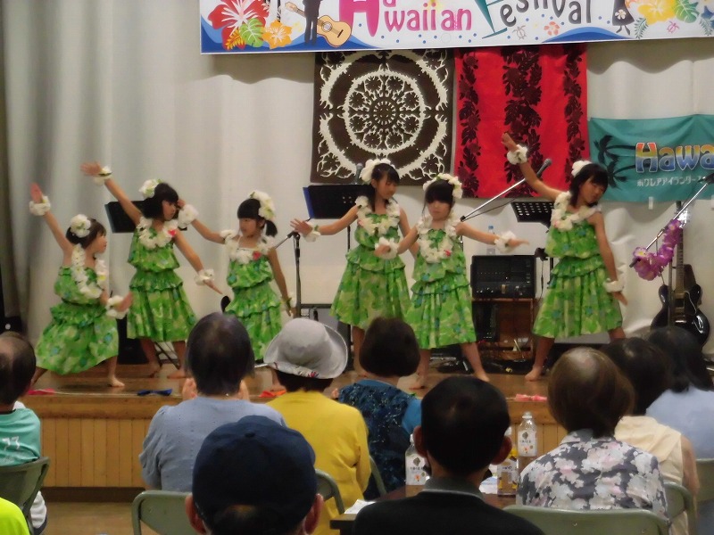 緑の衣装と白い花のレイを身につけた子供たちが、ステージ上でハワイアンダンスを披露している様子を、観客席から撮影した写真