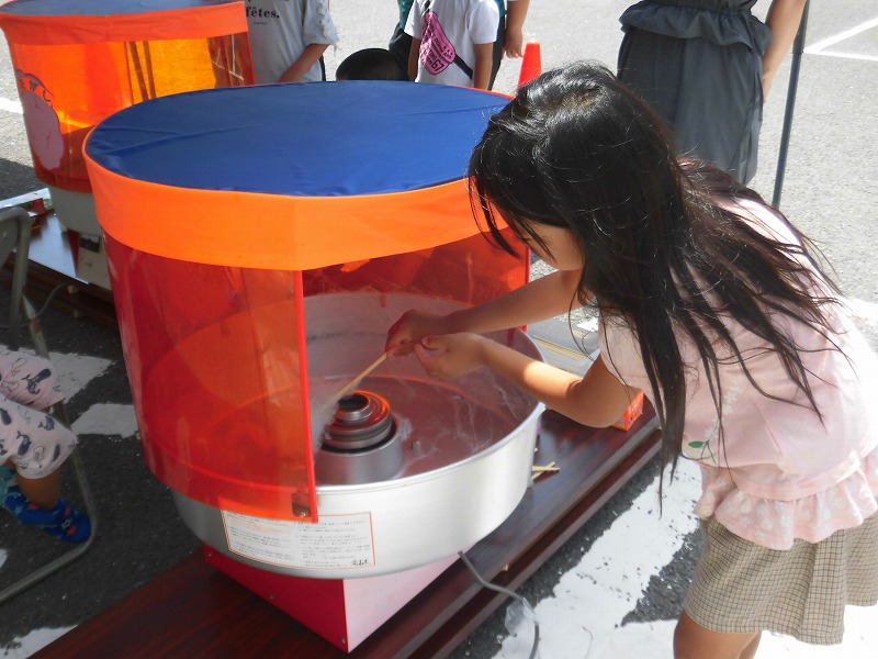 綿菓子製造機を使って、子供が木製の棒を機械の回転部分にあててふわふわの綿菓子を作っている写真