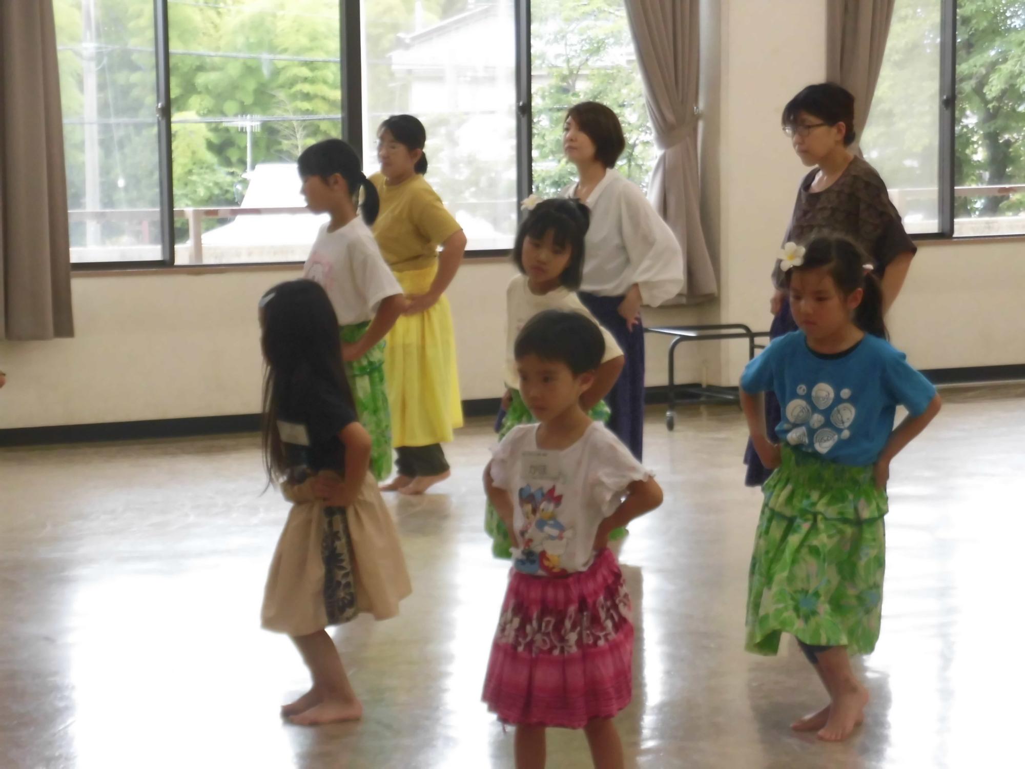 子どもから大人までの参加者が教室の中でフラダンスの練習をしており、それぞれがスカートを着用して手を腰にあてて真剣に竹内利枝さんの指導を受けている様子の写