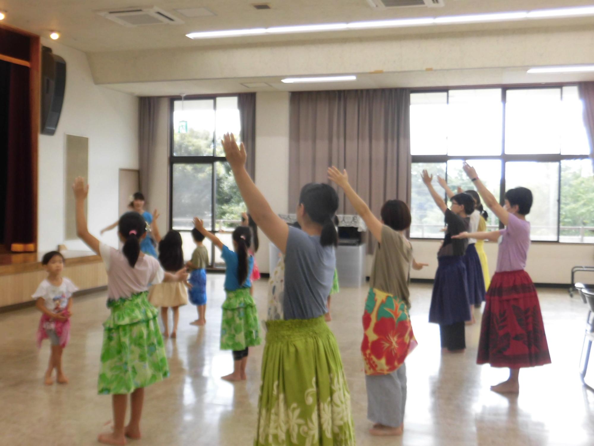 地域の多目的ホールで子どもから大人までの参加者が円になって竹内利枝さんの指導を受けながら、カラフルなスカートを身に着けて両手を高く上げるフラダンスの基本動作を練習している様子の写真