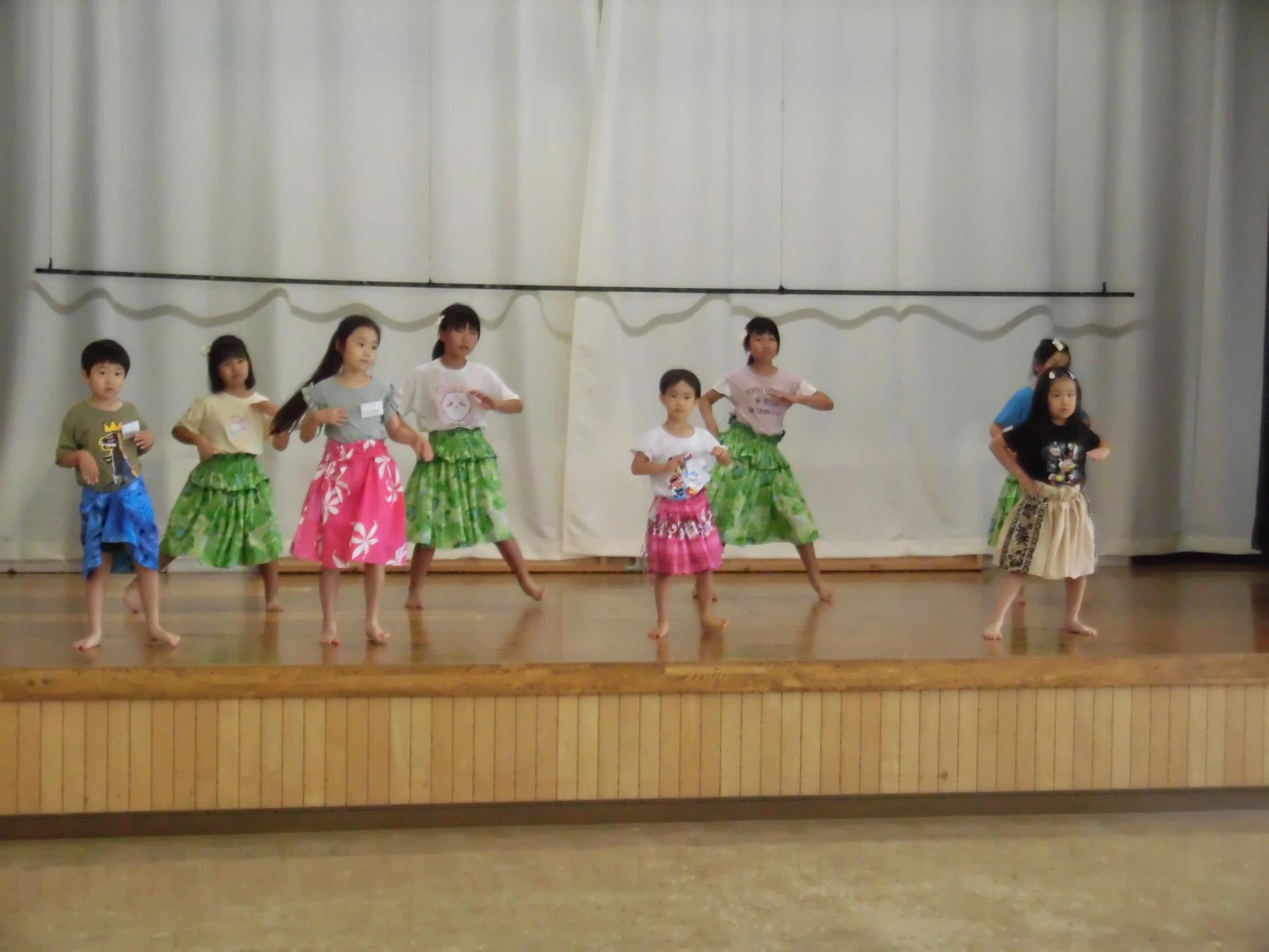 舞台の上でカラフルなパウスカートを身に着けた子どもたちが横一列に並び、手を腰にあてながらフラダンスの発表に向けた練習を真剣に行っている様子の写真