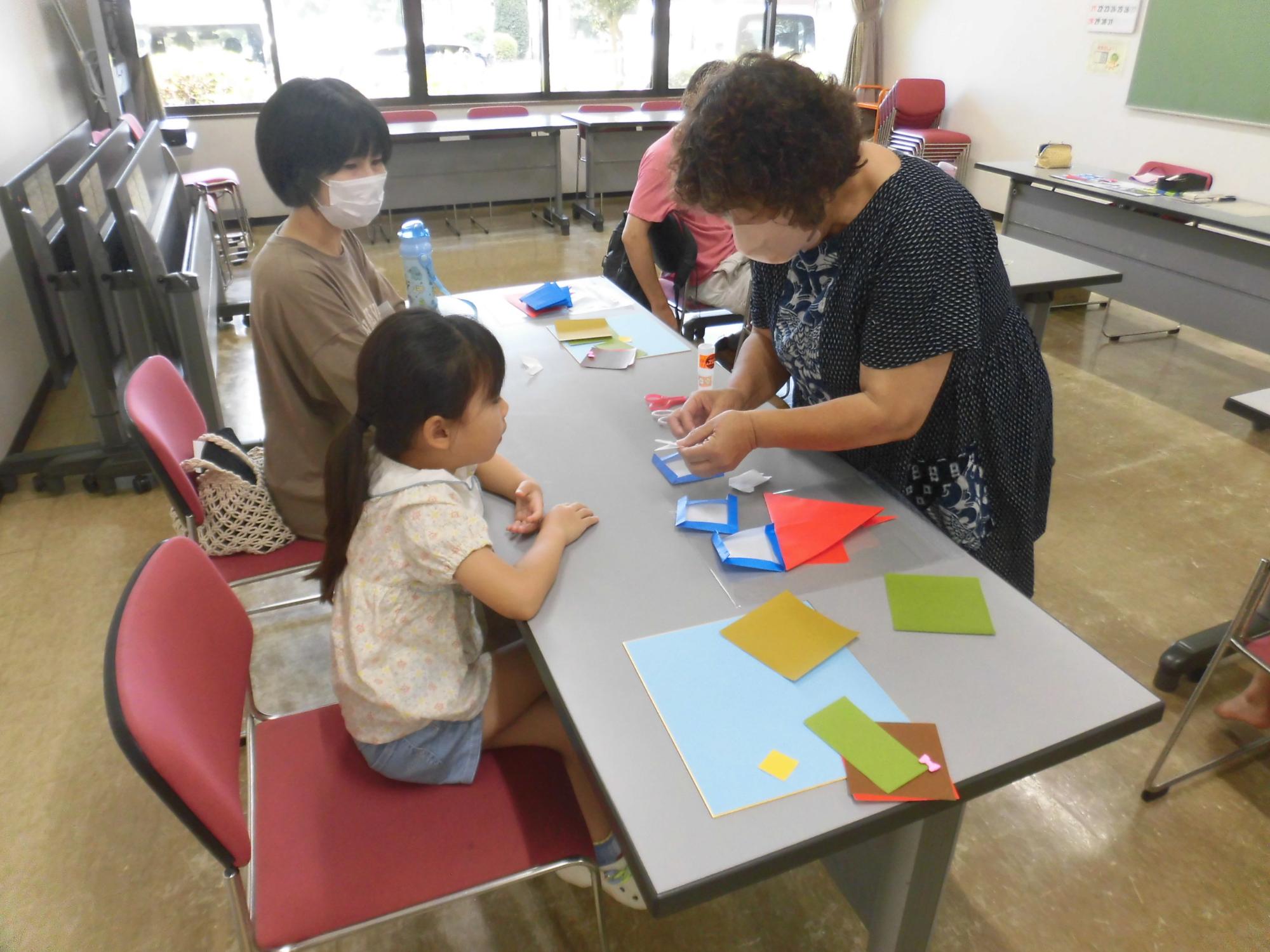 室内で女性と女の子がテーブルを囲んで座っており、山口みどりさんが折り紙のような紙を使って女の子に工作を教えている場面を写した写真
