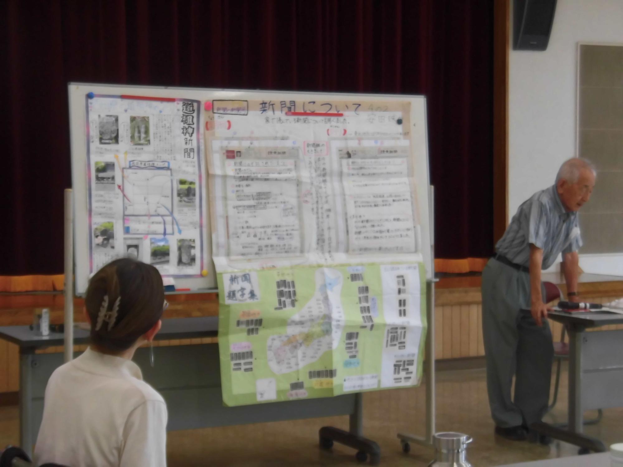 男性がホワイトボードに貼り付けられた新聞や地図について、座っている女性に向かって説明している様子を捉えた写真