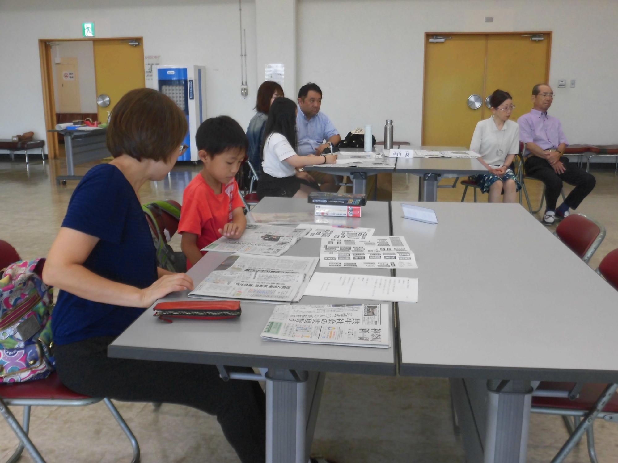 テーブルを囲んで複数の人々が座っており、手前では母親と子供が新聞を広げて熱心に読み、奥でも他の参加者が新聞を読んでいる様子の写真