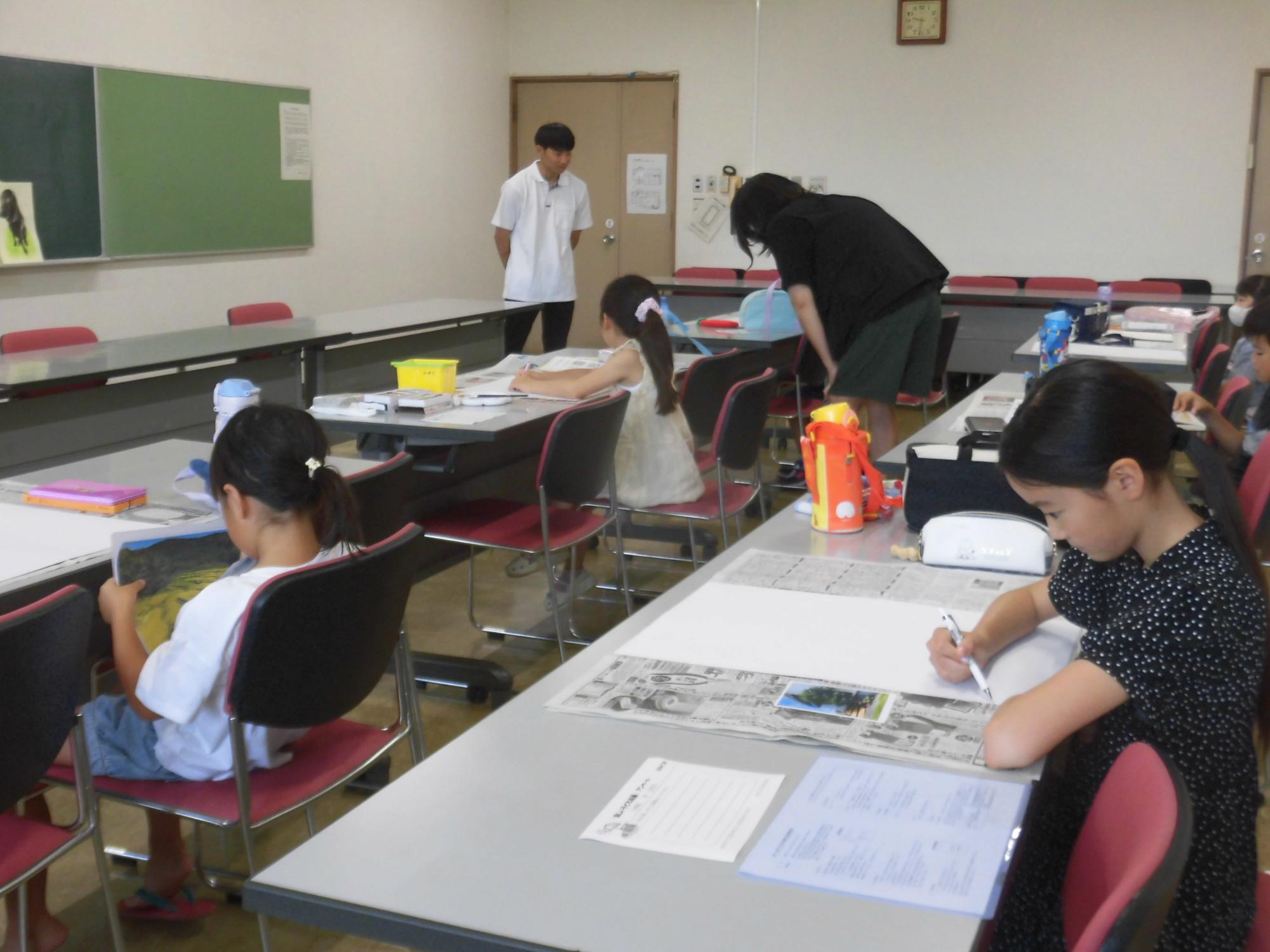 学習机が並んだ室内で、数人の子供が真剣な表情で座って何かを書き込んでおり、子ども絵画に取り組んでいる様子の写真